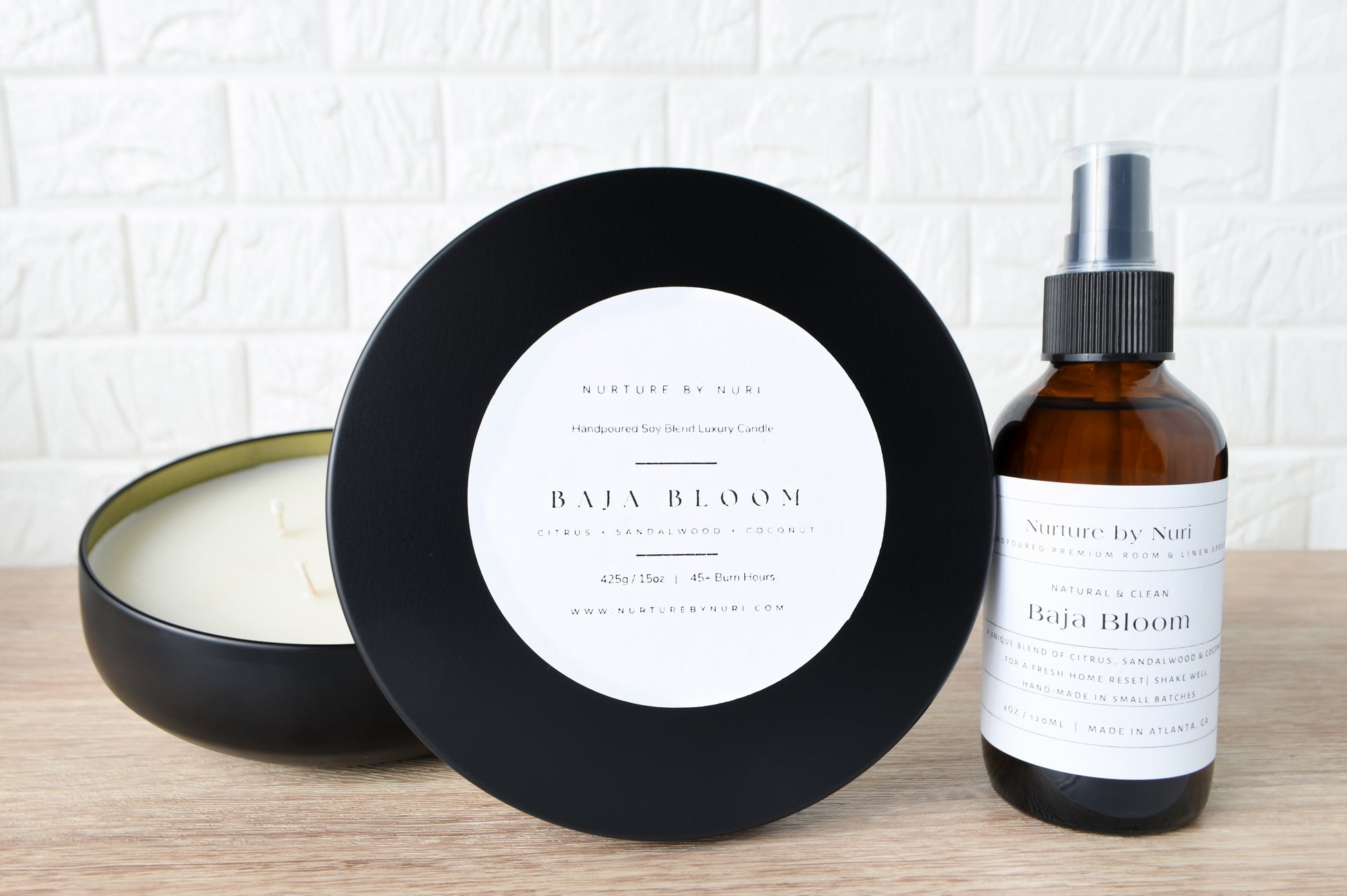 Nurture by Nuri Candle, black container, and bottle labeled 'Baja Bloom' on a wooden surface with a white brick wall background.